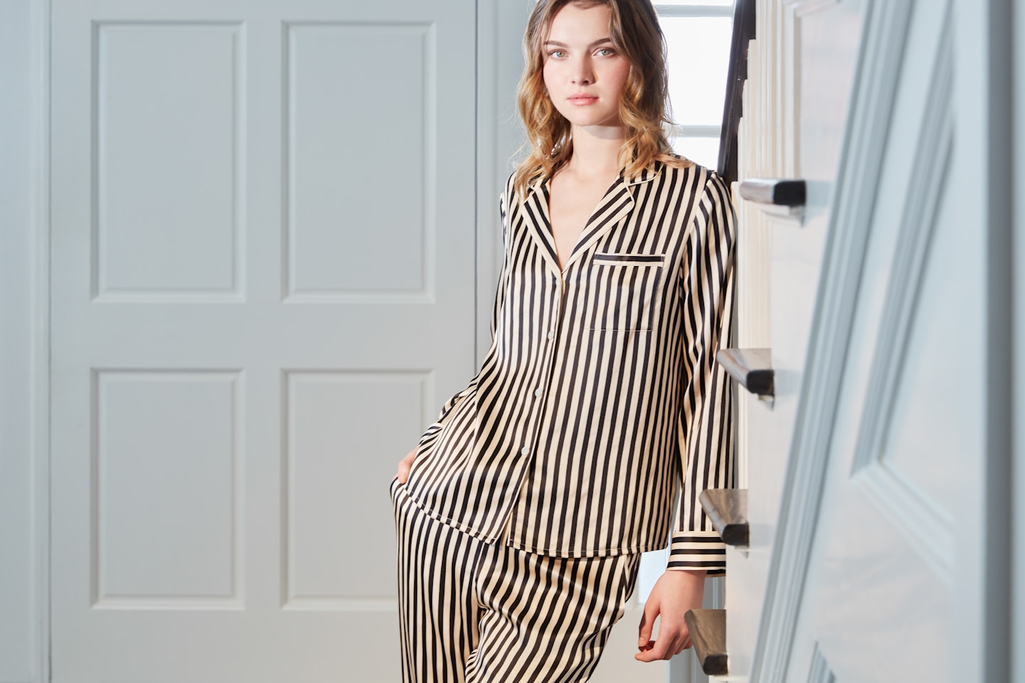 A woman leans by the staircase wearing a black and white striped MANITO pajama set, showcasing stylish sleepwear.